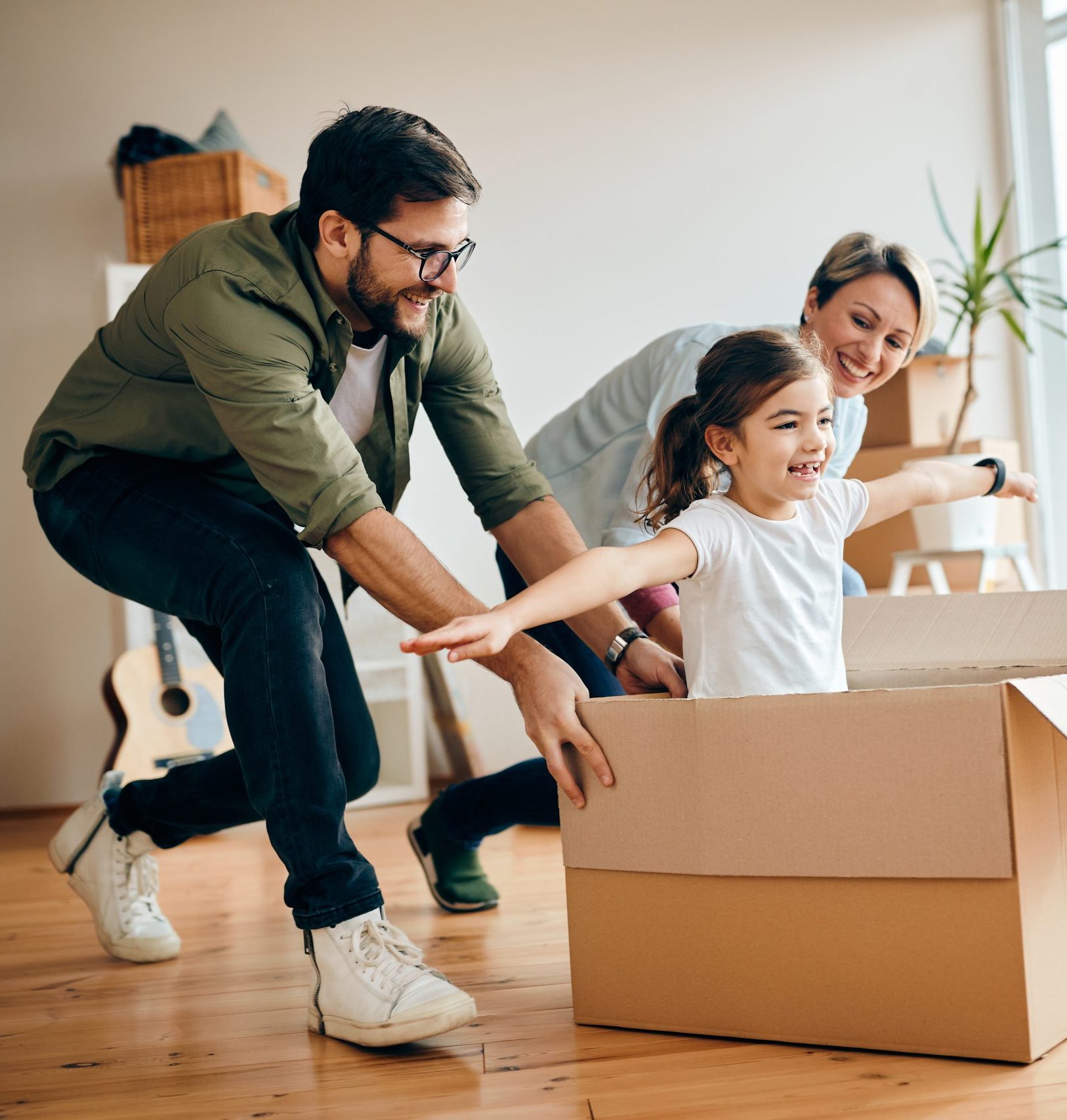 Happy little girl with arms outstretched having fun being pushed by her parents in carton box at new home